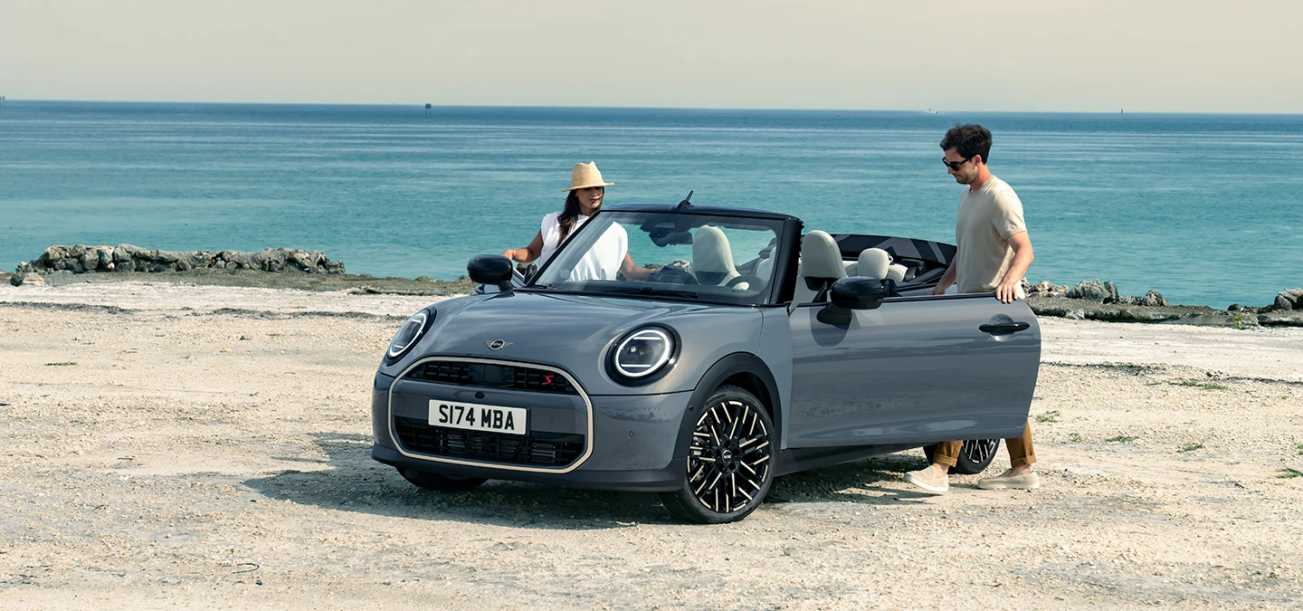 前左視角Copper Grey車色的MINI Cooper敞篷車停靠於海灘旁，旁有兩位乘客。